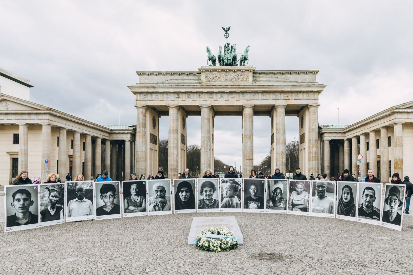 Vorne liegt das Mahnmal mit einem Kranz. Dahinter stehen Menschen in einem Halbkreis und halten sehr große Portraits hoch. Im Hintergrund das Brandenburger Tor.